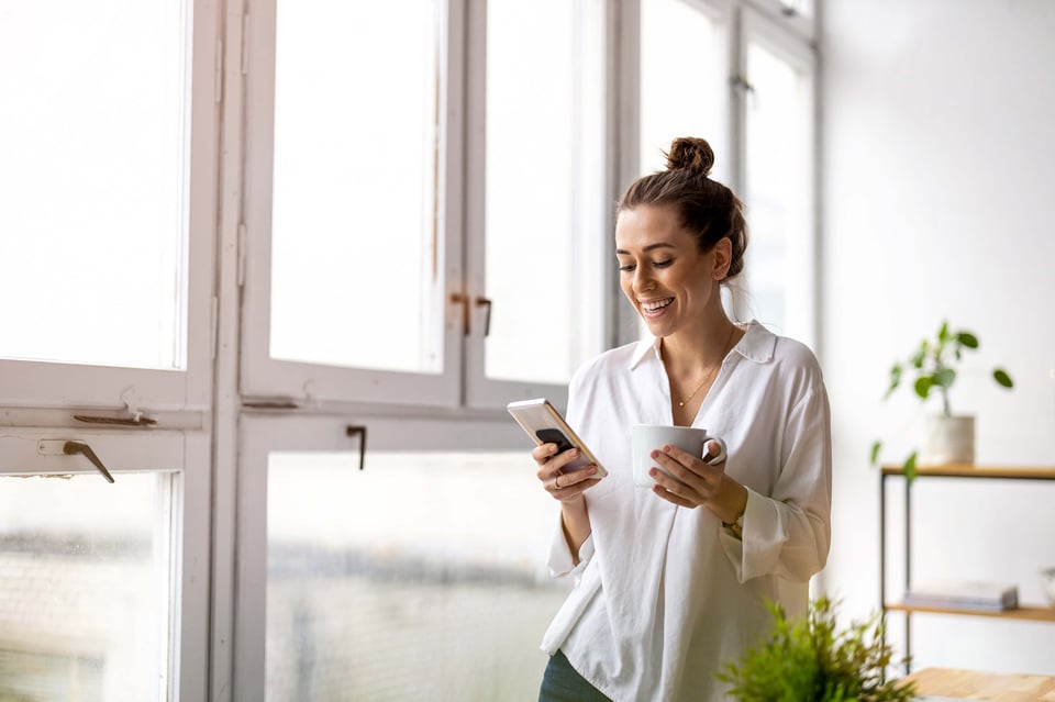 Kvinna som står hemma och tittar på mobilen med en kopp kaffe i den andra handen